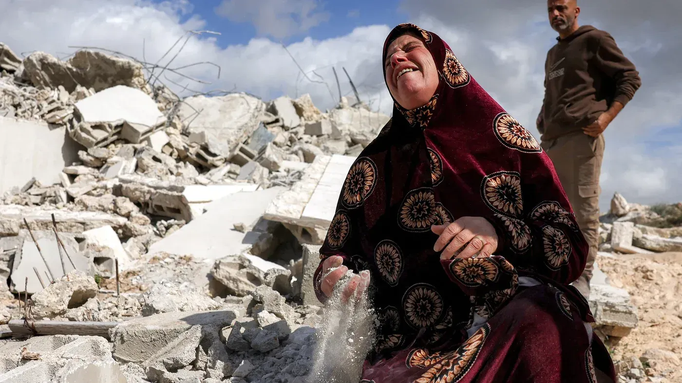 امرأة فلسطينية تجلس على أنقاض منزلها المهدوم، تعبر عن حزنها الشديد بينما يراقبها رجل خلفها، في سياق التهجير القسري في الضفة الغربية.