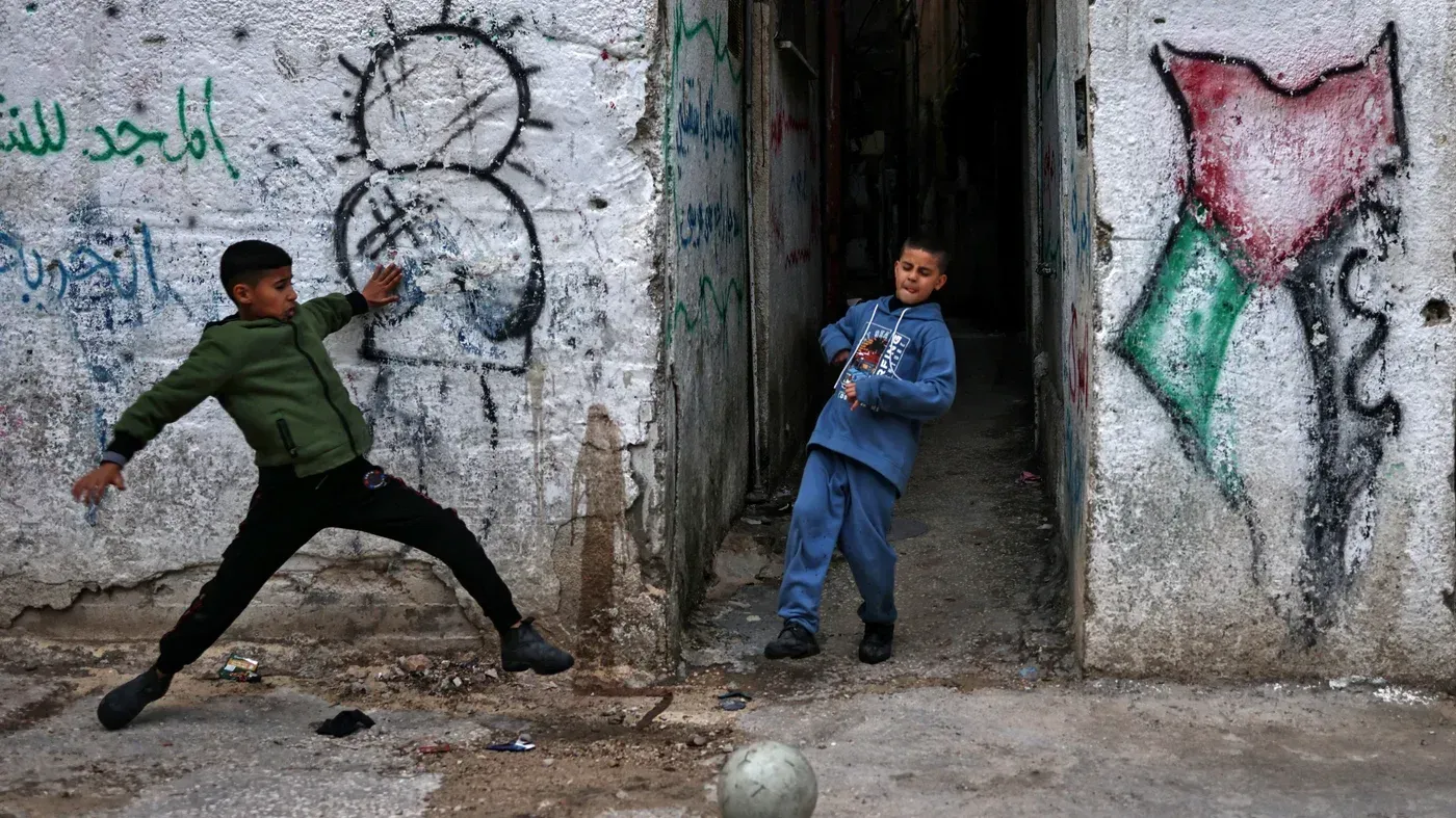طفلان فلسطينيان يلعبان في زقاق ضيق، مع جدران مزينة برسوم تعبر عن الهوية الفلسطينية، تعكس واقع الحياة في الضفة الغربية.