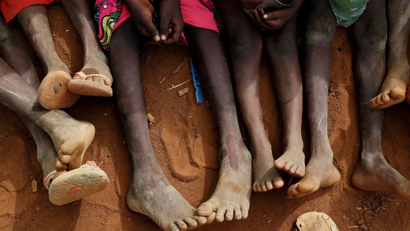 أرجل أطفال ملطخة بالتراب، تجلس في صف على الأرض، تعكس معاناة الأطفال في السودان وسط النزاع المستمر والاعتداءات.