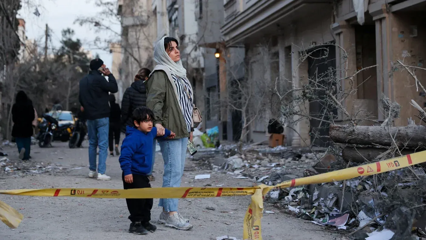 امرأة وطفل يقفان أمام منطقة مدمرة بعد الهجمات، مع وجود أشخاص آخرين في الخلفية، تشير إلى تأثير الصراع في المنطقة.