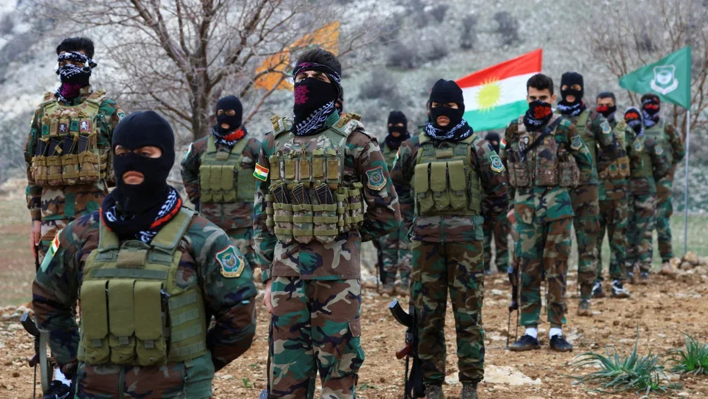 مقاتلون من البيشمركة الكردية يرتدون زيًا عسكريًا ويستعدون لمواجهة التهديدات الإيرانية، مع الأعلام الكردية في الخلفية.