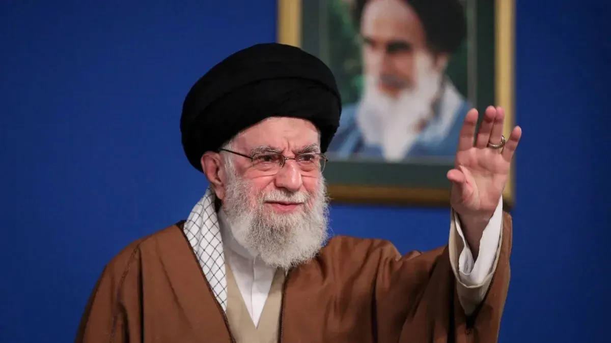 خامنئي، المرشد الأعلى الإيراني، يلوح بيده خلال خطاب، مع صورة الإمام الخميني في الخلفية، في سياق التوترات حول البرنامج النووي الإيراني.