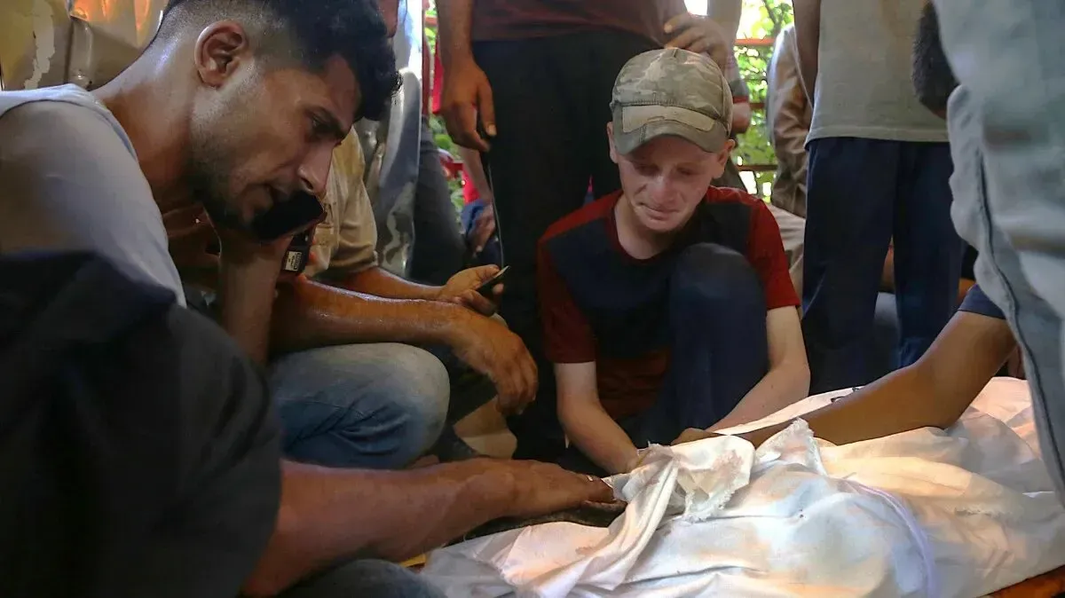 مشهد يعكس الحزن والألم، حيث يجتمع عدد من الأشخاص حول جثة مغطاة، مع تعبيرات وجهية تظهر الصدمة والفقد، في سياق الوضع الإنساني في غزة.