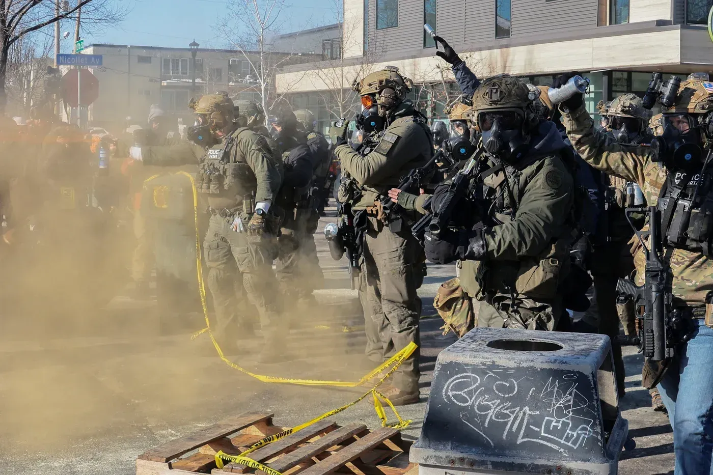 عناصر من الشرطة يرتدون زيًا عسكريًا ويستخدمون معدات تكتيكية خلال مواجهة في مينيابوليس، وسط غبار كثيف وأجواء متوترة.