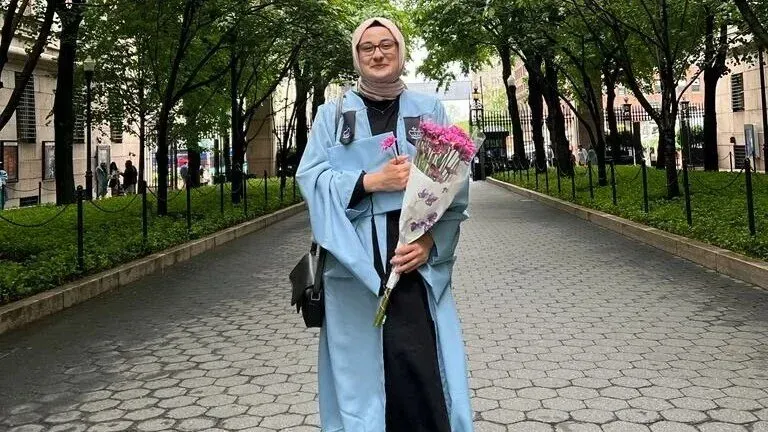 طالبة ترتدي زي التخرج وتحمل باقة من الزهور، تقف في ممر محاط بالأشجار، تعبير عن الفخر والنجاح في دراستها.