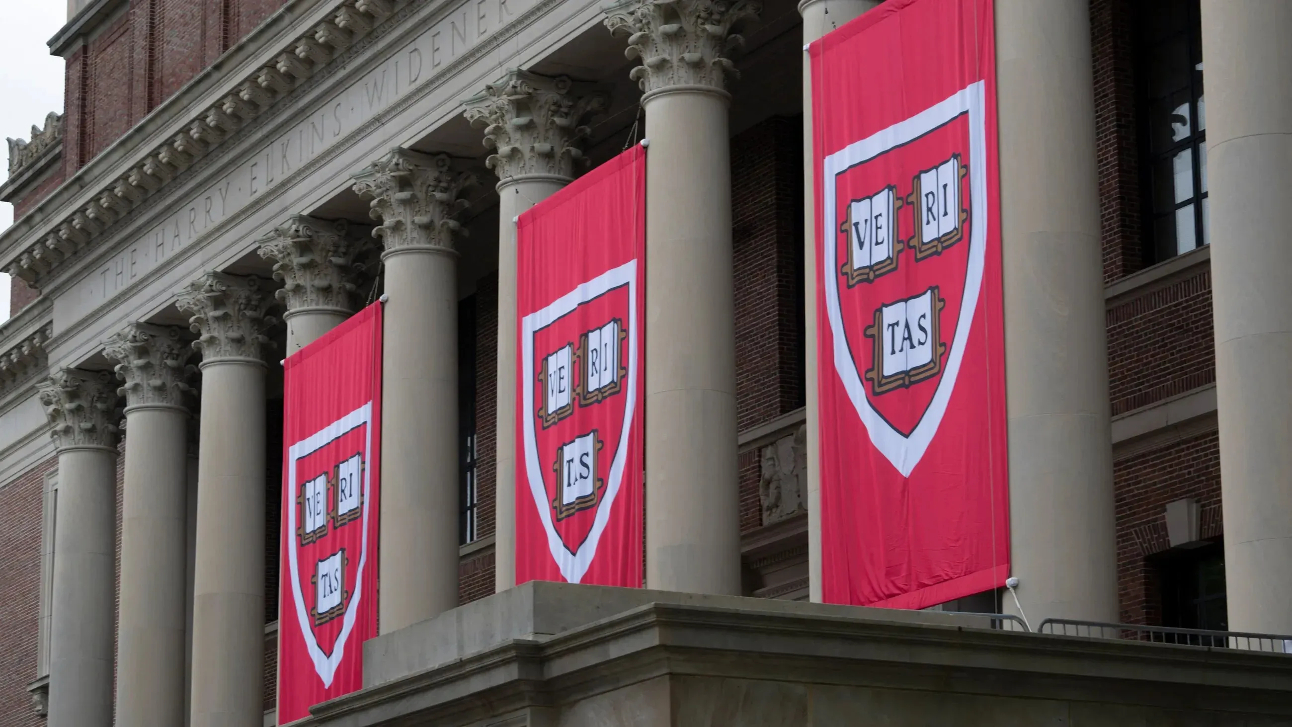 لافتات حمراء تحمل شعار جامعة هارفارد "VERITAS" معلقة على أعمدة المبنى، في سياق النزاع القانوني حول تسجيل الطلاب الدوليين.