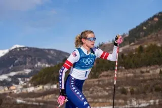 رياضية أمريكية تتزلج في سباق التزلج الريفي على الثلج، مرتدية زي الأولمبياد، مع مناظر جبلية في الخلفية.