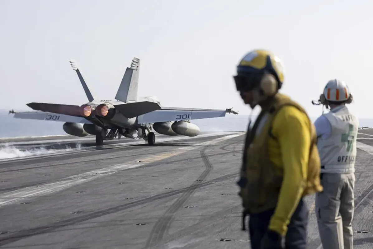 طائرة مقاتلة من طراز F/A-18 تقلع من حاملة طائرات، مع وجود أفراد عسكريين يرتدون زي العمل على سطح الحاملة، في سياق العمليات العسكرية ضد داعش.