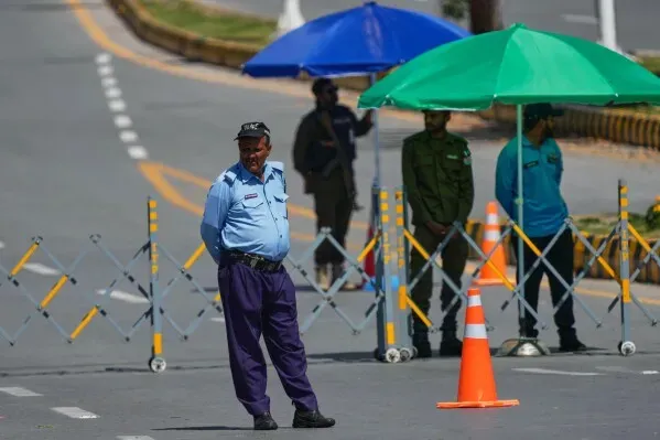 رجل أمن يقف بجانب حواجز مرورية في منطقة مفتوحة، مع مظلات ملونة في الخلفية، مما يعكس حالة من التأهب الأمني.