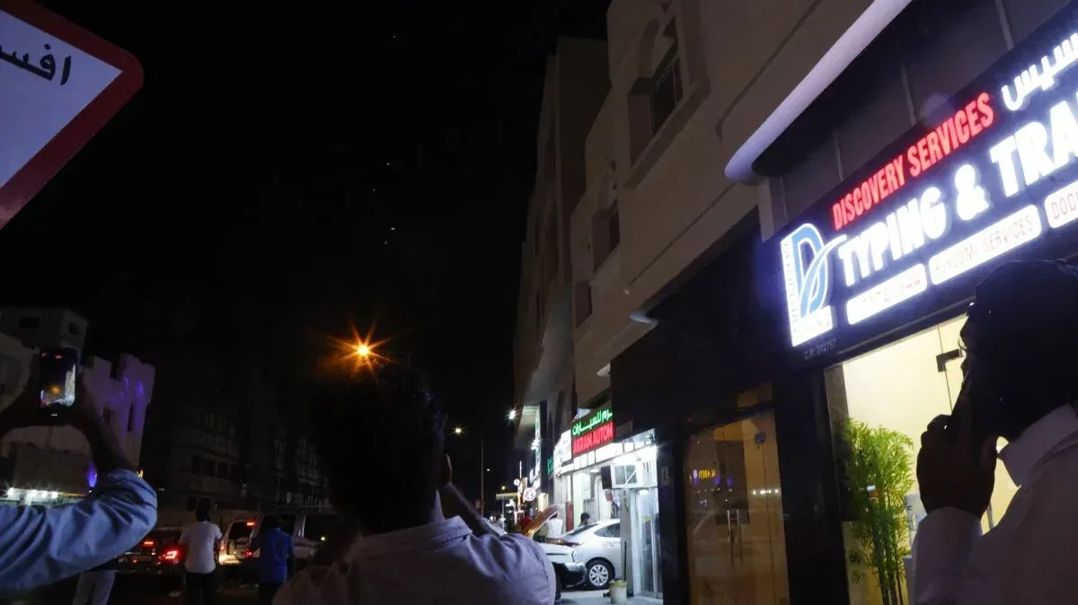 سكان في الشارع يلتقطون صورًا بهواتفهم تحت الأضواء الليلية، مع لافتة تشير إلى خدمات الطباعة في الخلفية، مما يعكس أجواء التوتر في المنطقة.