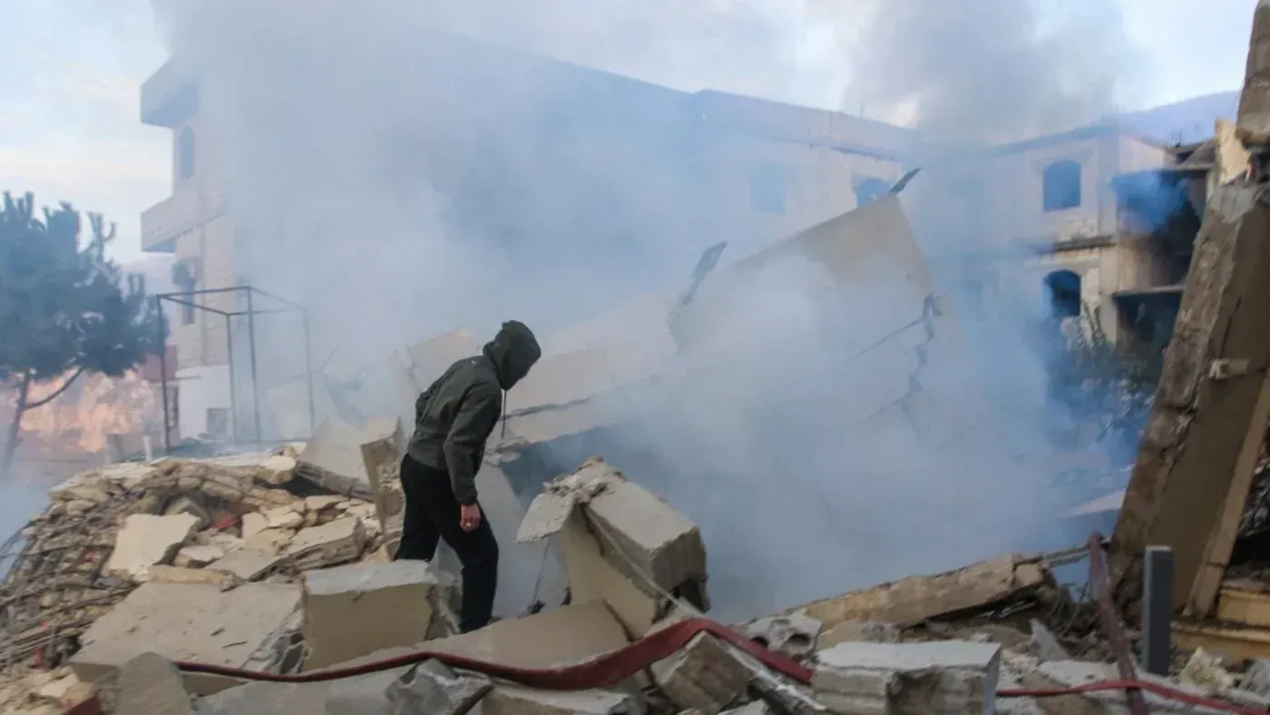 شخص يرتدي سترة واقية يتجول بين الأنقاض والدخان في منطقة تضررت من الغارات الجوية الإسرائيلية في لبنان، مما يعكس تصاعد التوترات السياسية.