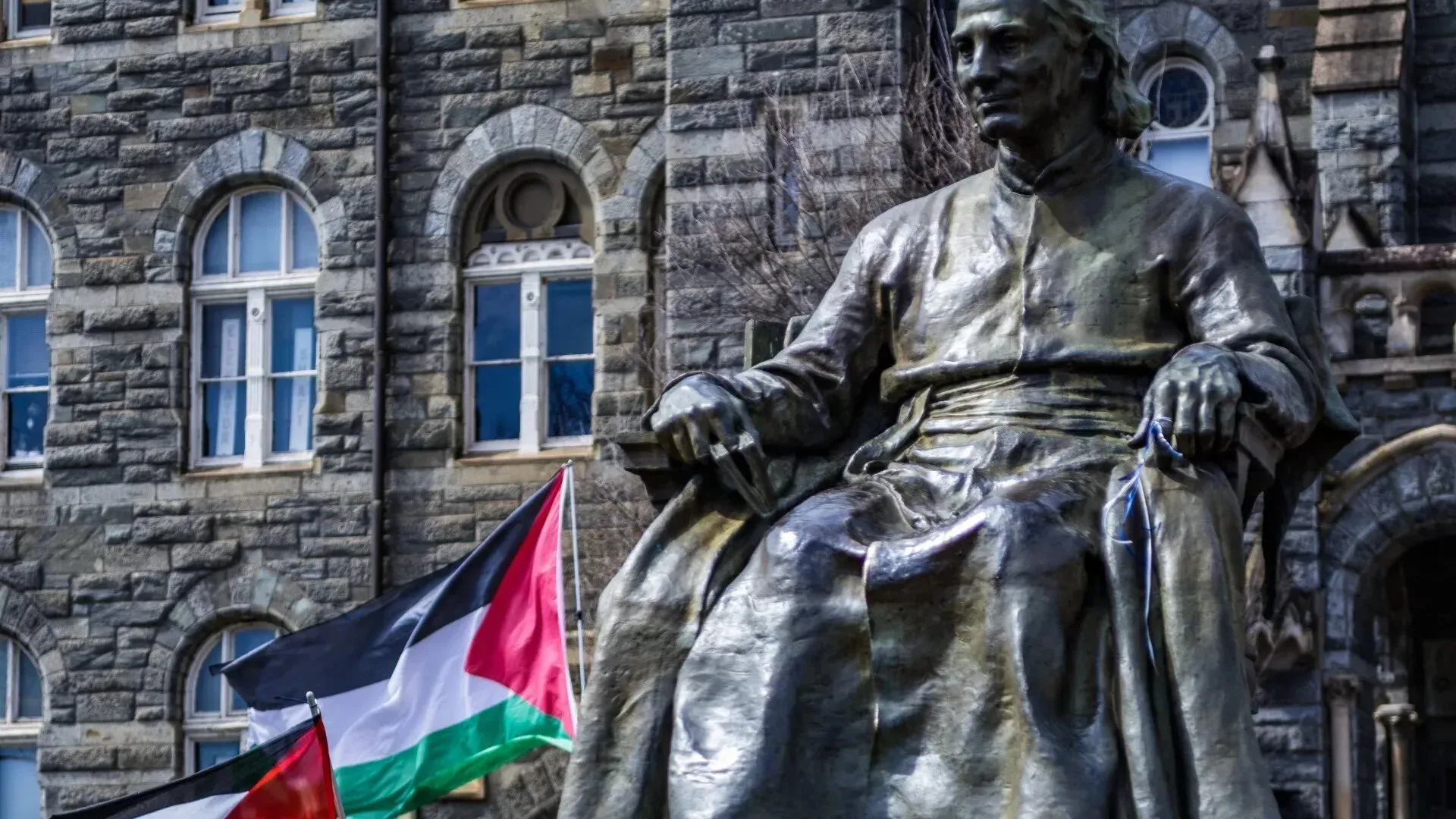 تمثال برونزي لشخصية تاريخية جالس أمام مبنى حجري، مع وجود علم فلسطين يرفرف بجانبه، مما يعكس سياق القضايا المتعلقة بفلسطين.