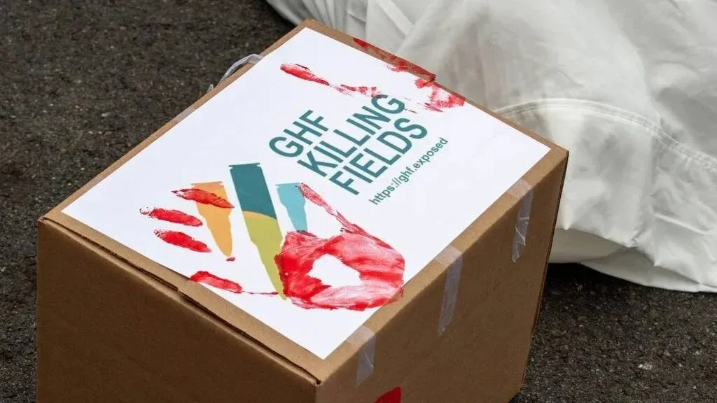 صندوق غذائي يحمل علامة "GHF KILLING FIELDS" مع آثار دماء، يرمز إلى الضحايا الفلسطينيين في غزة خلال احتجاج ضد المؤسسة الإنسانية.