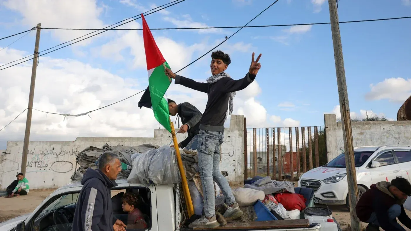 طفل فلسطيني يحمل علم فلسطين واقفاً على سيارة محملة بالأمتعة، بينما تظهر خلفه مجموعة من الناس والأبنية في غزة.