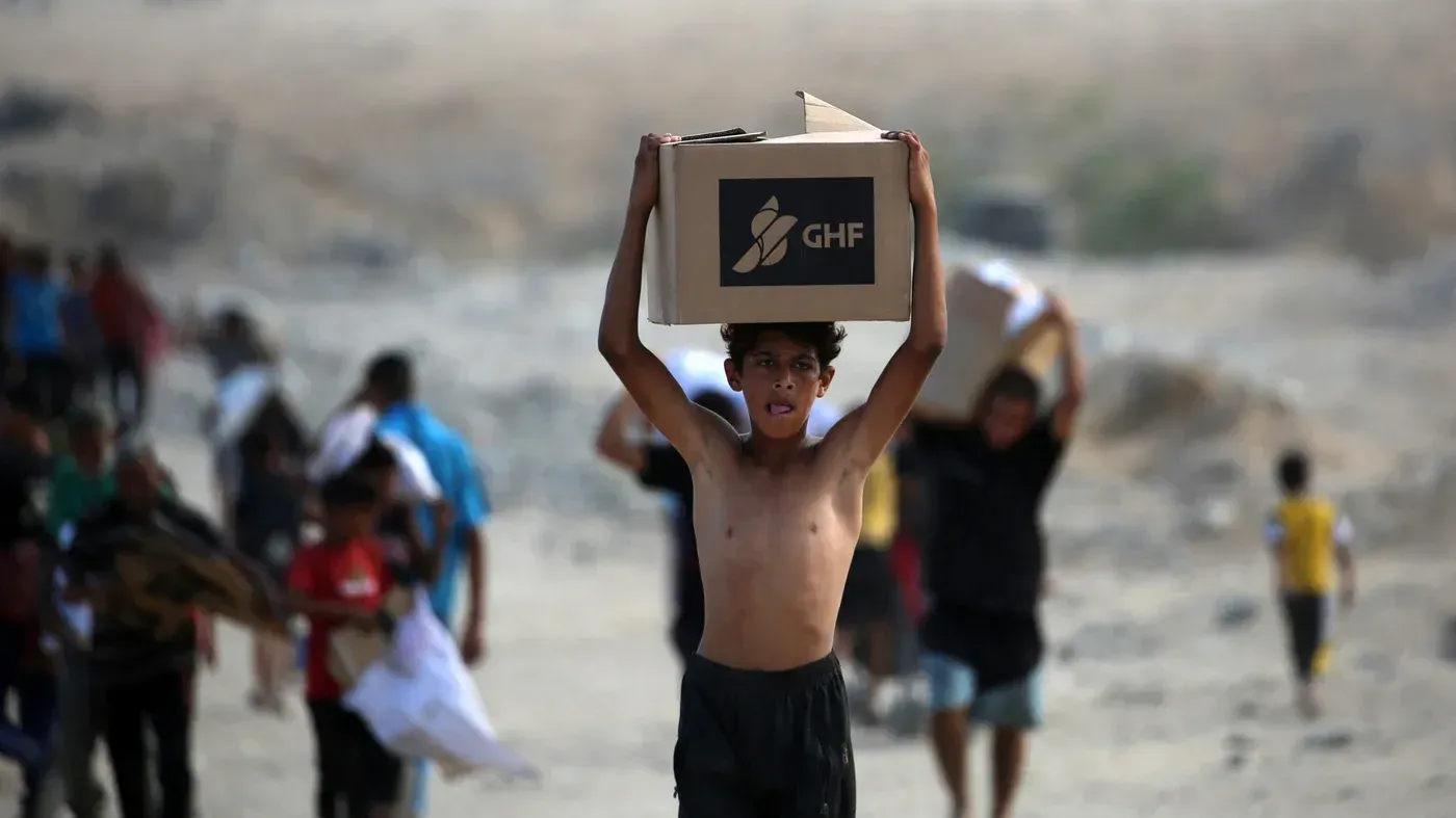 طفل يحمل صندوق مساعدات من مؤسسة GHF وسط مجموعة من الأشخاص في غزة، مما يعكس الأوضاع الإنسانية الصعبة في المنطقة.