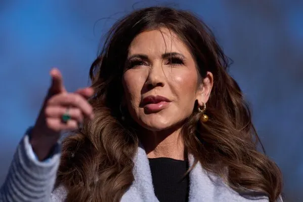 وزيرة الأمن الداخلي الأمريكية كريستي نويم تتحدث في حدث عام، مشيرة بيدها، مع خلفية سماء زرقاء.