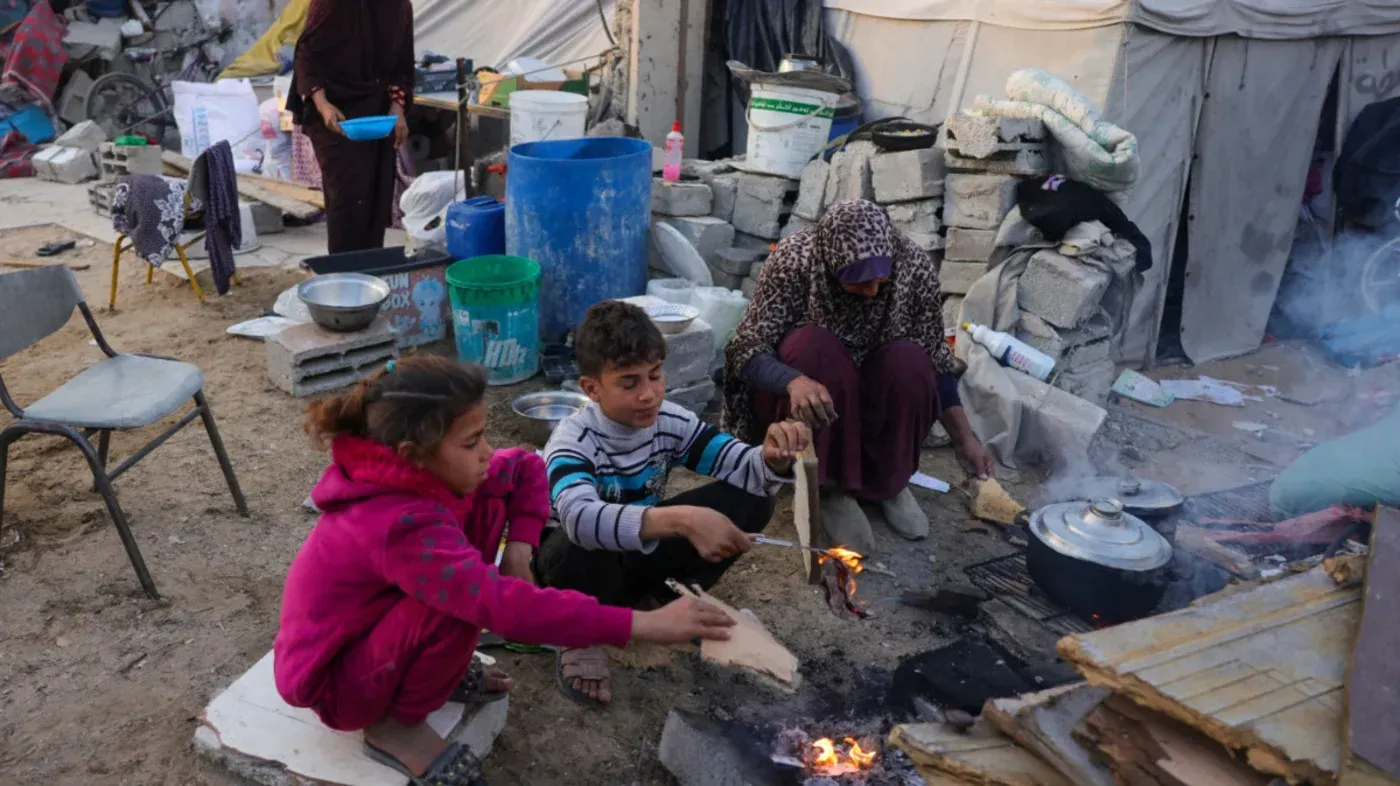عائلة فلسطينية تطهو الطعام في مخيم مؤقت، محاطة بأدوات منزلية بسيطة، تعبيرًا عن الحياة اليومية في ظل ظروف صعبة بعد الحرب.