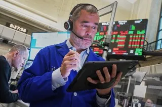تاجر في بورصة الأسهم الأمريكية يستخدم جهاز لوحي، مع شاشات تعرض مؤشرات السوق في الخلفية، في ظل ارتفاع الأسهم بعد بيانات سوق العمل.