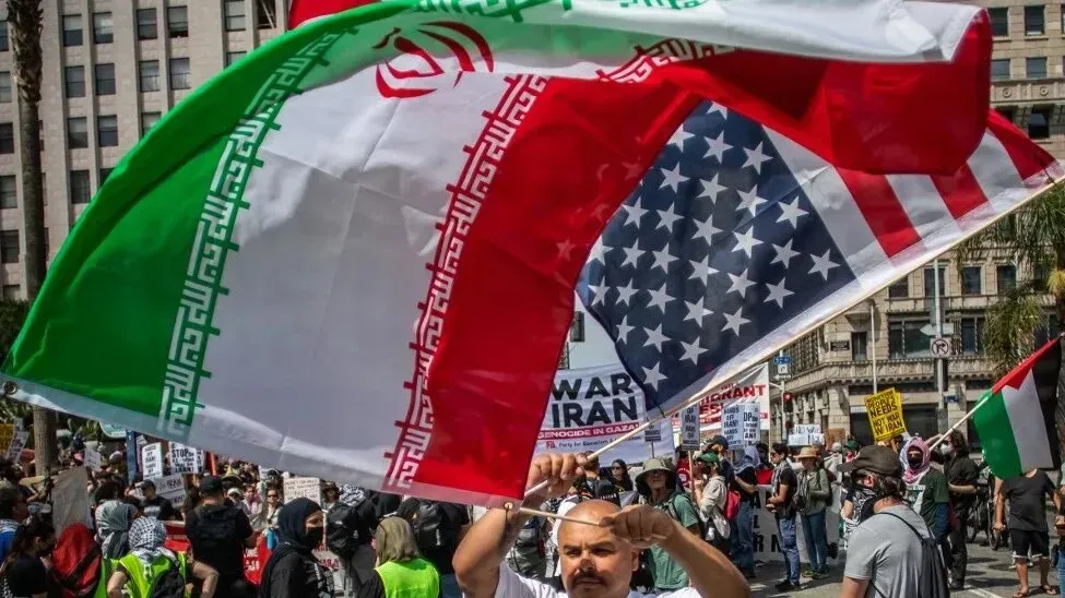 متظاهر يحمل علمي إيران والولايات المتحدة في احتجاج ضد الضربات الأمريكية على المواقع النووية الإيرانية، مع وجود حشود في الخلفية.