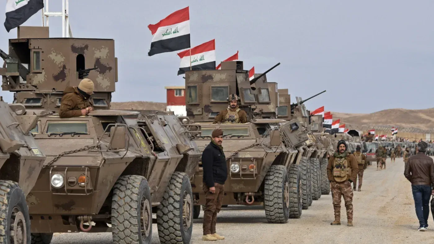 عرض لموكب عسكري عراقي يتضمن مركبات مدرعة وجنودًا، مع أعلام العراق ترفرف في الخلفية، في سياق التوترات السياسية الحالية.