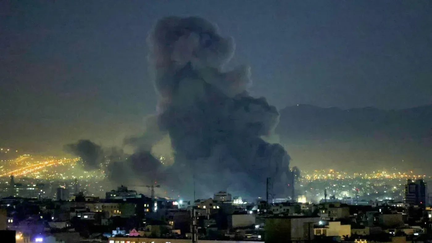 دخان كثيف يتصاعد من منطقة حضرية في الليل، مع أضواء المدينة في الخلفية، مما يعكس تداعيات الهجوم الأمريكي الإسرائيلي على إيران.