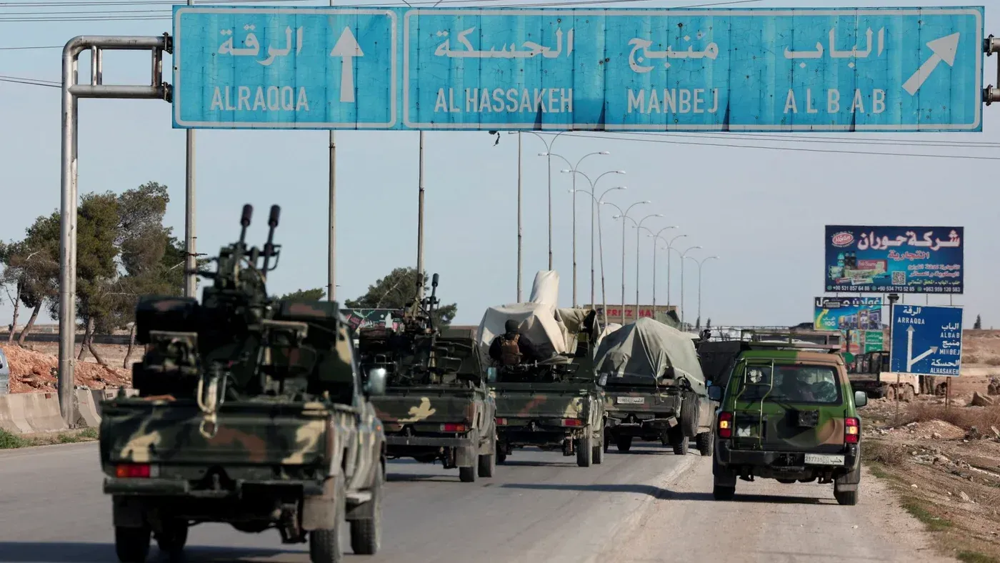 قافلة من المركبات العسكرية تسير على طريق في سوريا، مع لافتات تشير إلى الاتجاهات نحو الرقة والحسكة ومنبج.