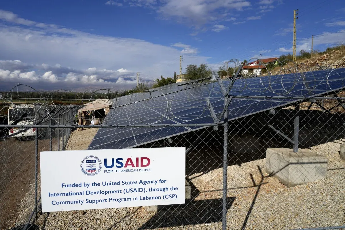 لوحة إعلانات لوكالة USAID توضح تمويل مشروع دعم المجتمع في لبنان، مع وجود ألواح شمسية وخلفية طبيعية.
