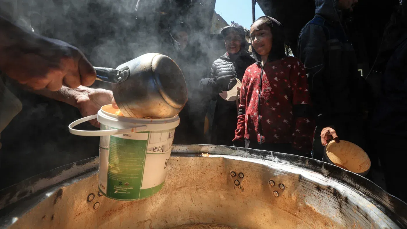 تظهر الصورة شخصاً يسكب الطعام من وعاء كبير في ساحة عامة، حيث يتجمع عدد من الأشخاص، بينهم أطفال، في انتظار المساعدة الغذائية وسط أزمة إنسانية في غزة.