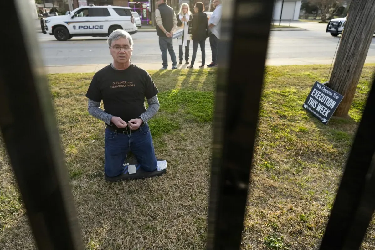 رجل يت kneel في حديقة أمام سجن ساوث كارولينا، مع لافتة تعبر عن المعارضة للإعدام، بينما يقف مجموعة من الأشخاص في الخلفية.