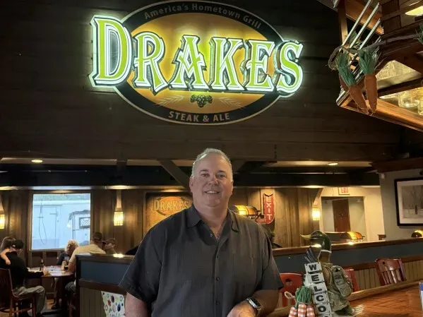 رجل يقف مبتسمًا أمام لافتة مطعم "Drakes Steak & Ale"، مع أجواء داخلية مريحة وخلفية تضم تفاصيل من المطعم.
