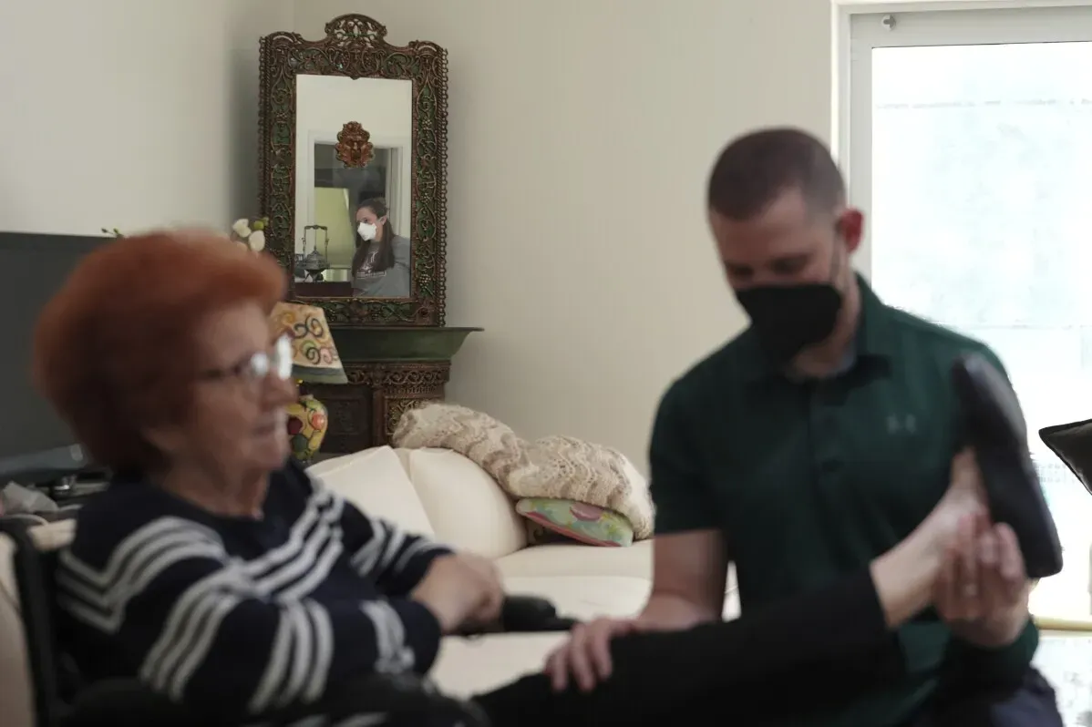 امرأة مسنّة تجلس على أريكة بينما يقوم معالج يرتدي قناعًا بفحص قدمها، مع انعكاس شخص آخر يرتدي قناعًا في المرآة خلفهم.