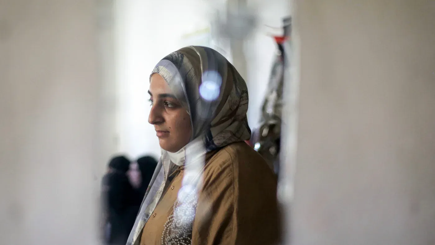 شابة فلسطينية ترتدي الحجاب تقف بجانب نافذة في غزة، تعبيرها يعكس الصبر والأمل وسط ظروف صعبة للتعليم والحياة.