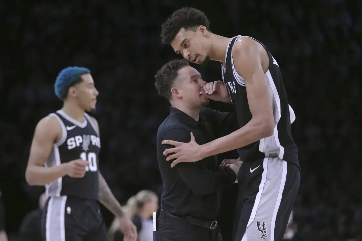يظهر في الصورة فيكتور ويمبانياما، نجم كرة السلة، يتفاعل مع أحد المدربين خلال مباراة في دوري NBA في باريس، وسط أجواء حماسية.