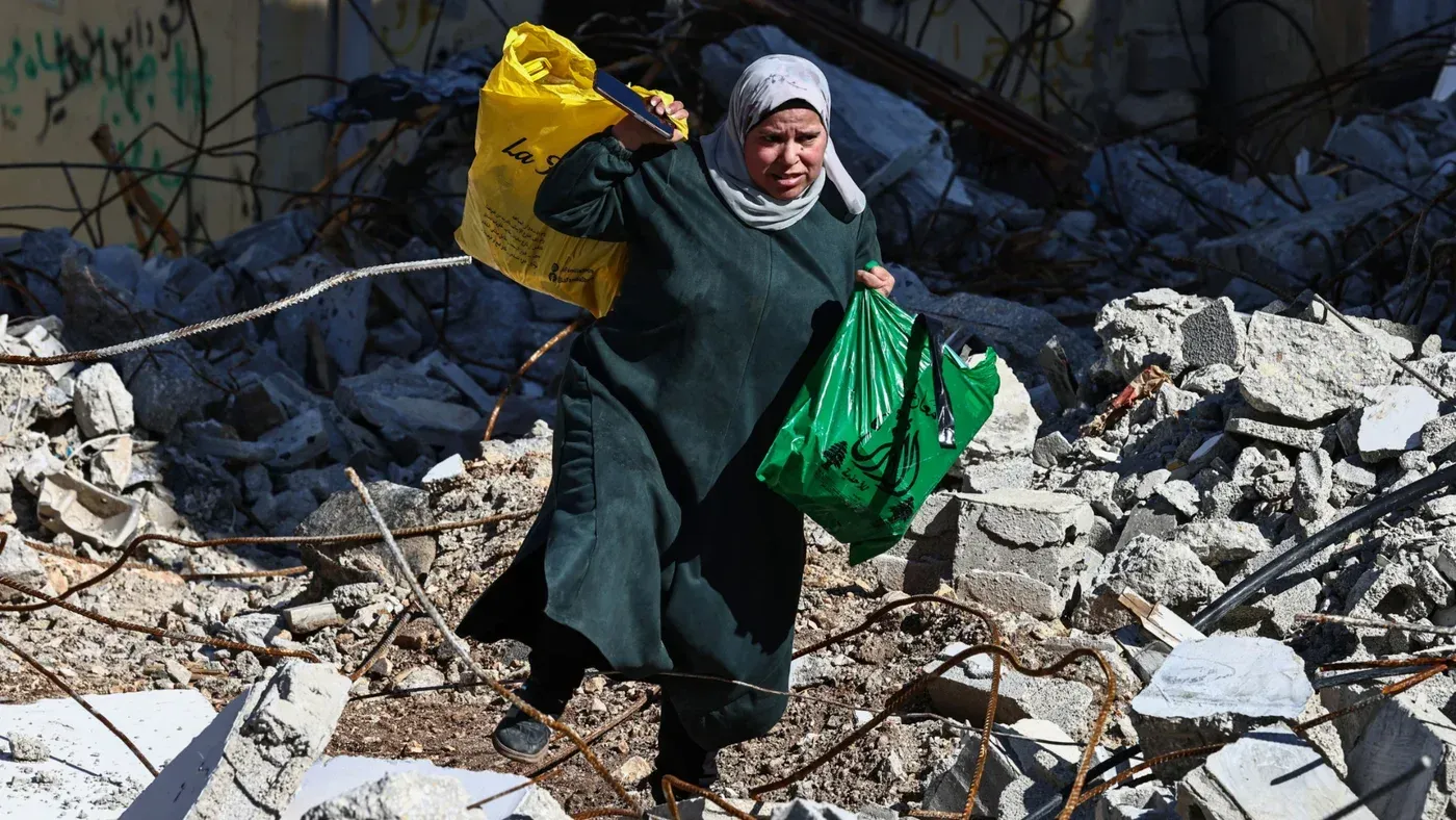 امرأة فلسطينية تحمل أكياسًا في منطقة مدمرة، تعكس معاناة النازحين خلال شهر رمضان في مخيم جنين.
