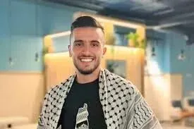 شاب فلسطيني مبتسم يرتدي كوفية، يظهر في بيئة داخلية مع تفاصيل خلفية بسيطة، تعكس الحياة اليومية في المجتمع الفلسطيني.