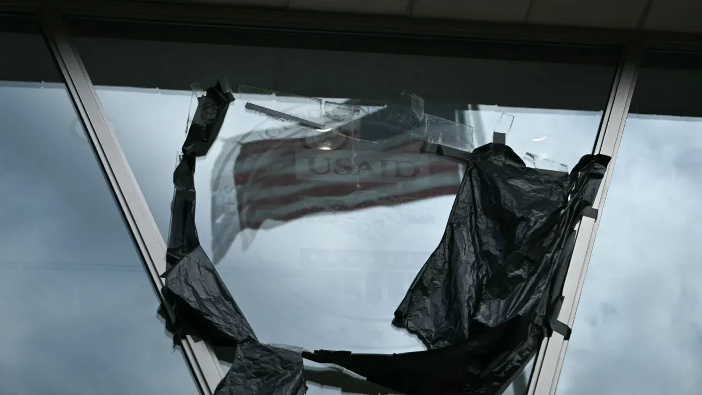شباك مكسور يظهر علم الوكالة الأمريكية للتنمية الدولية (USAID) مغطى بغطاء أسود، مما يعكس تأثير تخفيضات المساعدات الإنسانية.
