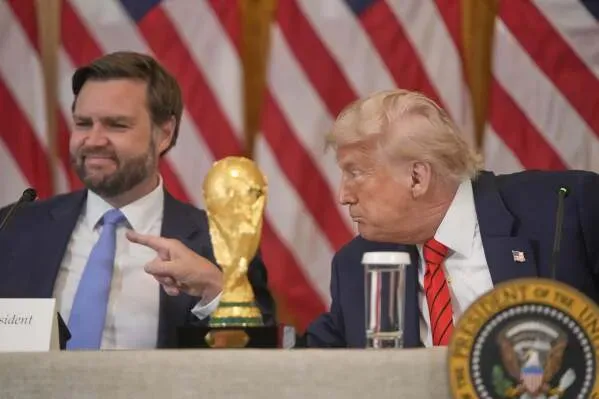 ترامب يشير إلى كأس العالم 2026 خلال مؤتمر صحفي، بينما يجلس بجانبه شخص آخر، مع خلفية من الأعلام الأمريكية.
