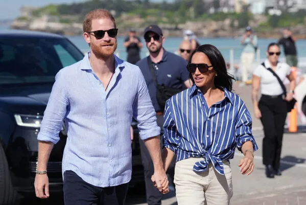 الأمير Harry وزوجته Meghan يمسكان أيديهما ويتجولان في مكان عام، مع أشخاص في الخلفية، في سياق عودتهما إلى بريطانيا بعد غياب طويل.