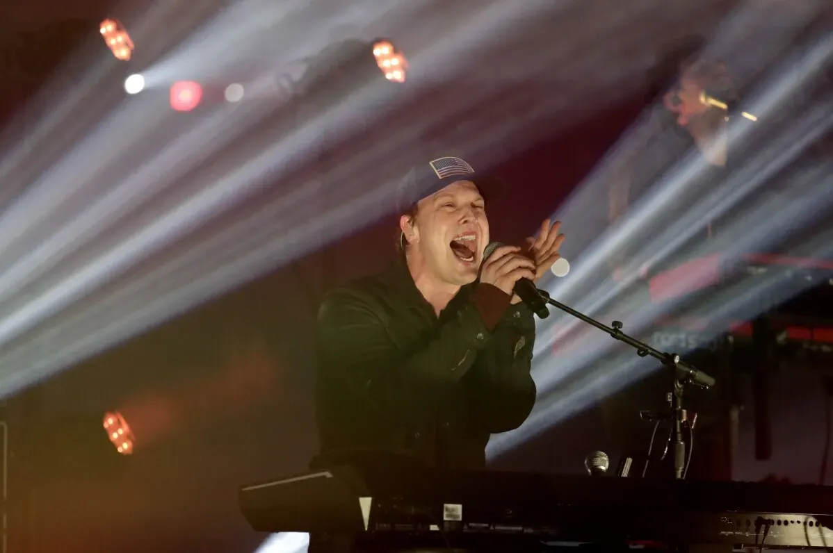 مغني يؤدي عرضًا حيويًا على المس stage، محاطًا بأضواء ساطعة، معبّرًا عن حماس كبير خلال حفل تنصيب ترامب.