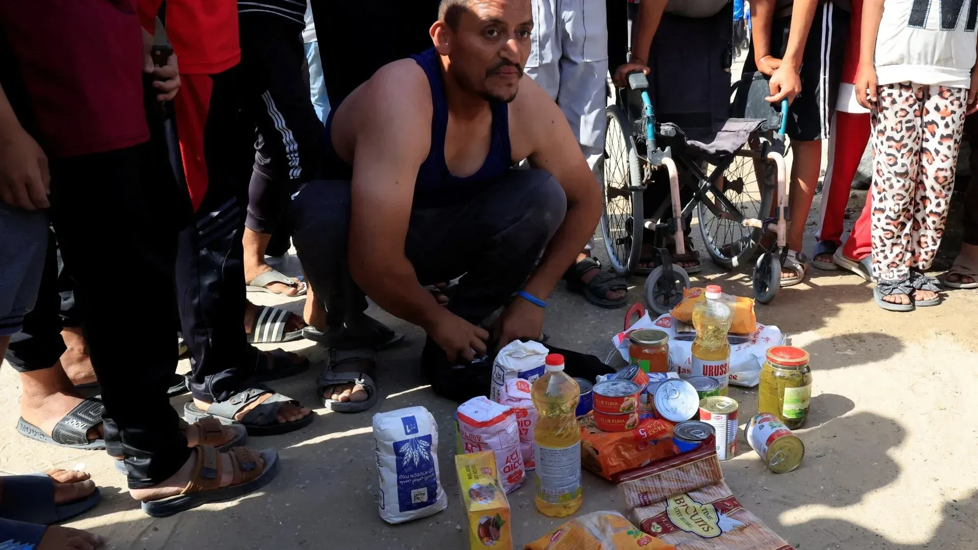 رجل فلسطيني يجلس على الأرض محاطًا بمجموعة من المواد الغذائية الأساسية مثل الزيت، الأرز، والدقيق، في سياق توزيع المساعدات في غزة.