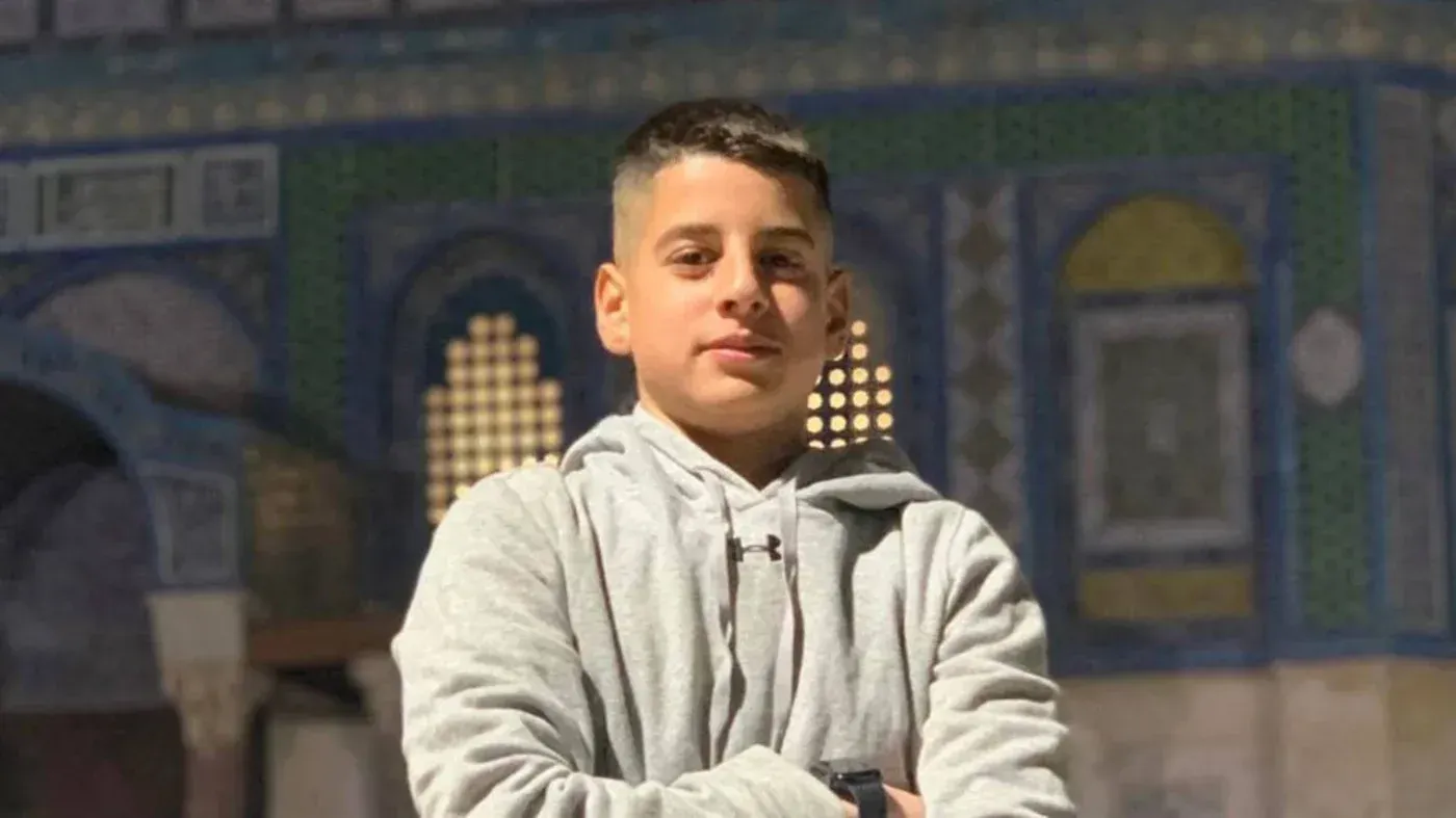 صورة لصبي فلسطيني يقف مبتسمًا في مكان تاريخي، مع خلفية معمارية مميزة، تمثل معاناة الأطفال الفلسطينيين المعتقلين.