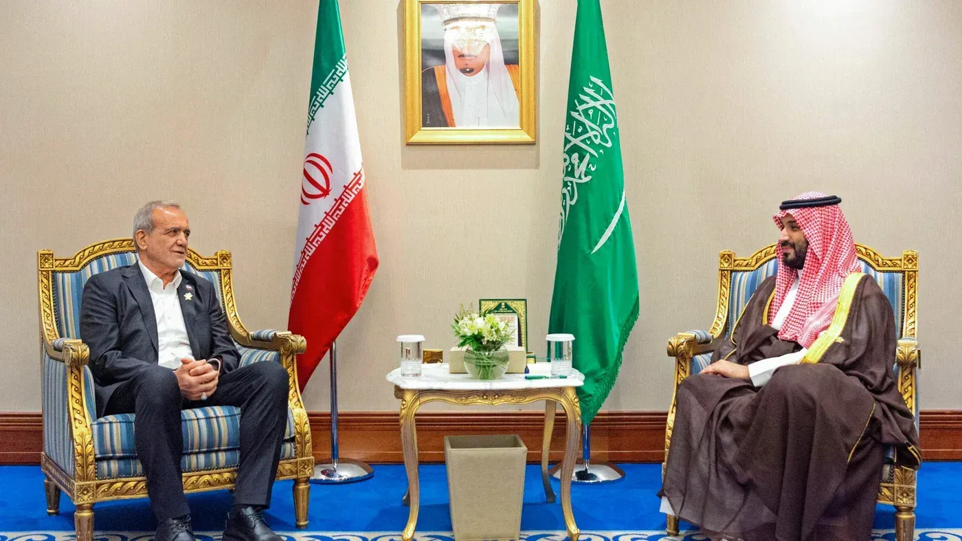 اجتماع بين مسؤول إيراني وولي العهد السعودي، مع العلميات الإيرانية والسعودية خلفهم، يعكس التوترات الإقليمية والتغيرات السياسية.