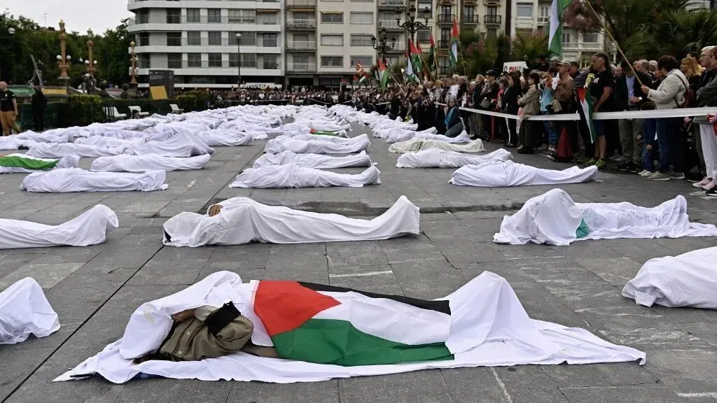 تظهر الصورة مجموعة من الجثث المغطاة بأقمشة بيضاء، مع أعلام فلسطينية، في احتفال تأبيني يرمز إلى الإبادة الجماعية للفلسطينيين.