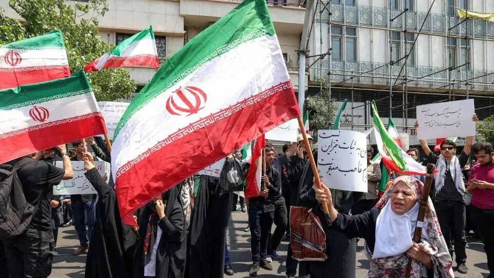 تظهر الصورة مظاهرة في إيران حيث يحمل المتظاهرون الأعلام الإيرانية، مع وجود لافتات تعبر عن مشاعر وطنية قوية.