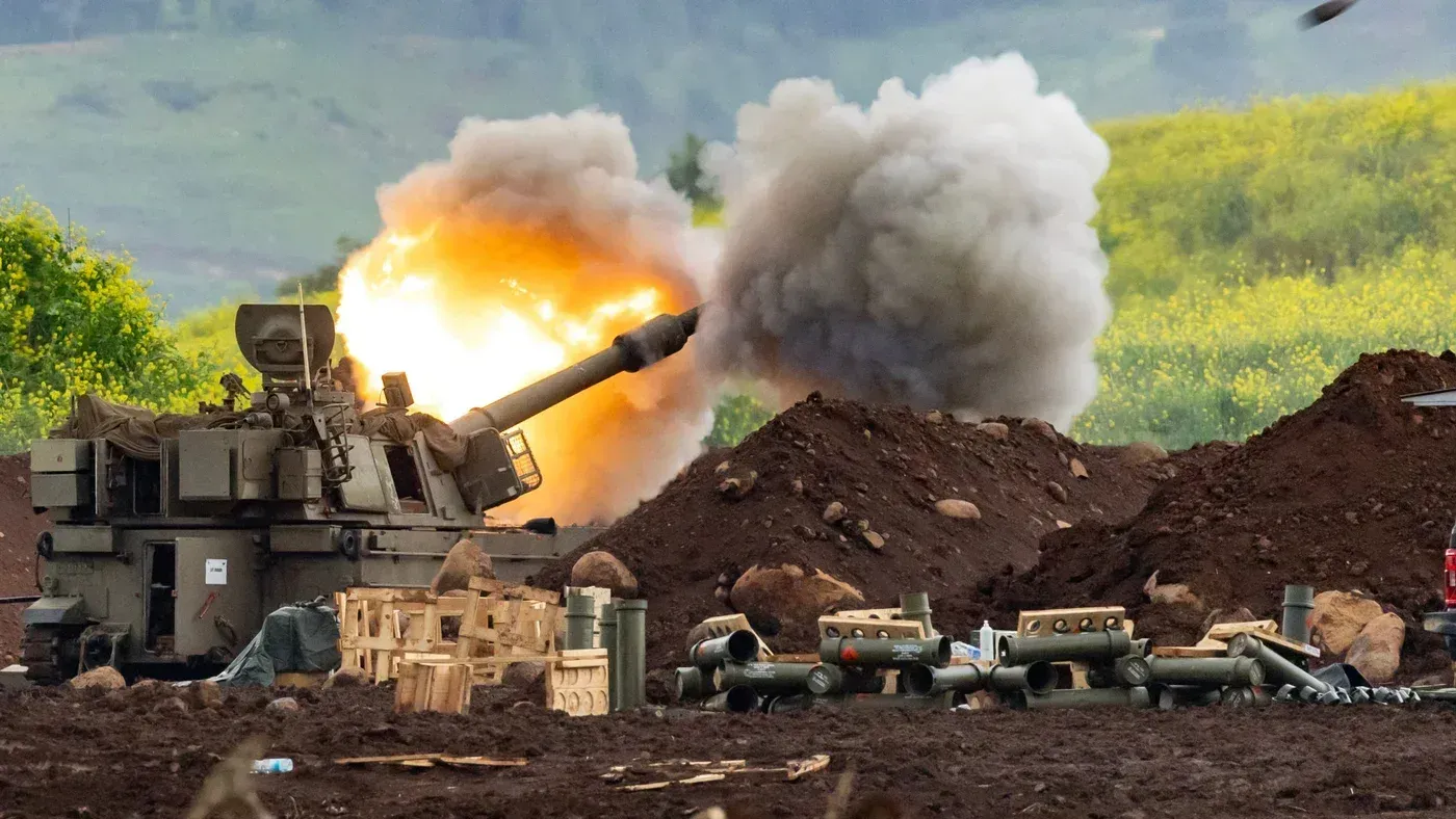 مدفع إسرائيلي يطلق قذيفة في منطقة زراعية، مع تصاعد الدخان والانفجار في الخلفية، مما يعكس تصاعد التوترات العسكرية في الشرق الأوسط.