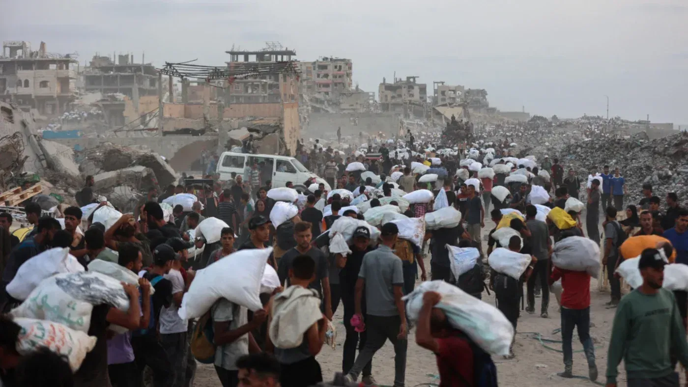 مواطنون فلسطينيون يحملون الأمتعة في منطقة مدمرة بغزة، وسط الدمار الناتج عن النزاع المستمر، مع تزايد المخاوف من المجاعة.