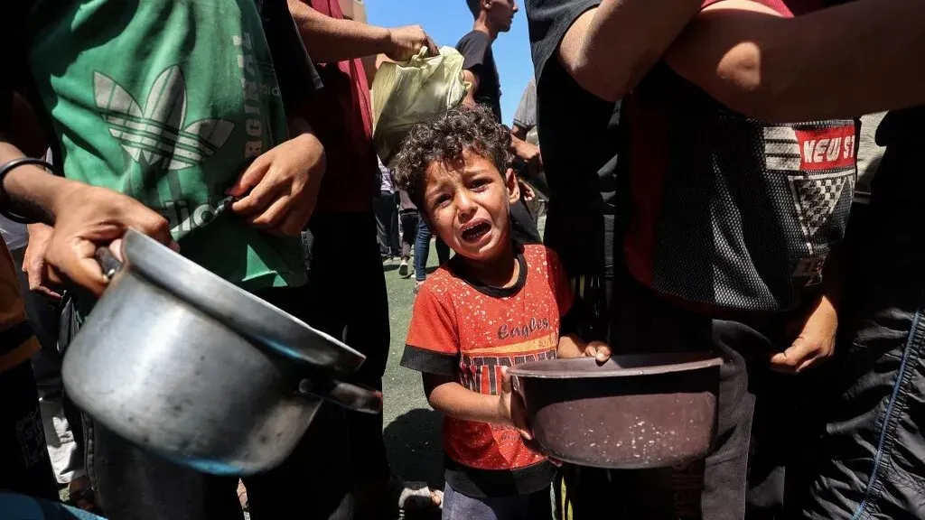 طفل فلسطيني يبكي في طابور للحصول على الطعام، محاط بأشخاص يحملون أكياسًا وأواني، مما يعكس معاناة السكان في غزة خلال الأزمة الإنسانية.