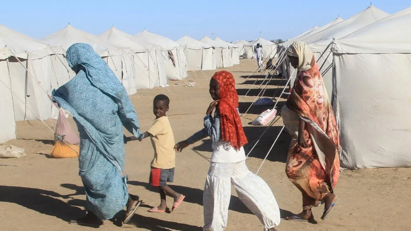 نساء وأطفال يسيرون في مخيم للنازحين في السودان، يعكس معاناتهم اليومية في ظل الأزمات المستمرة.