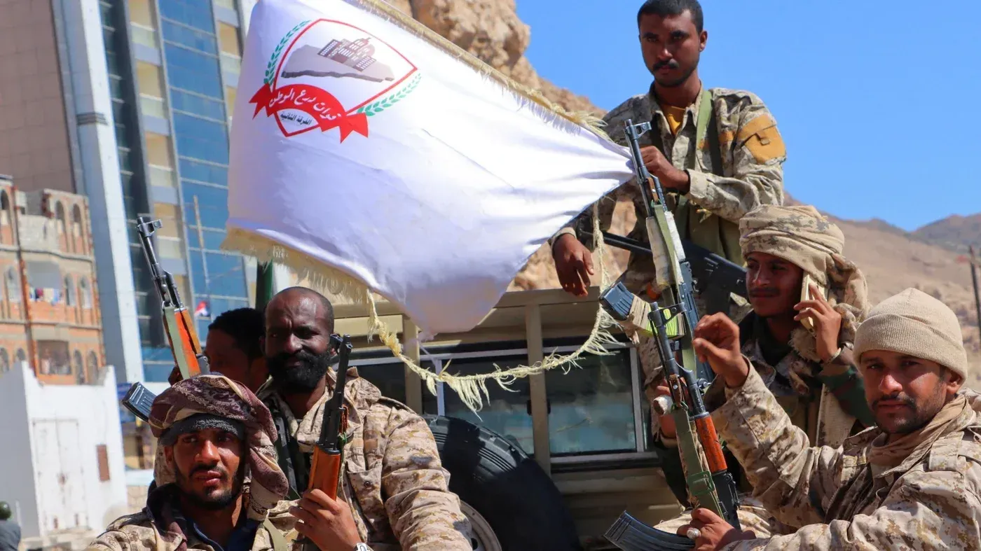 مقاتلون يمنيون يحملون أسلحة ويعرضون علمًا، في سياق الصراع المستمر في اليمن ودور الإمارات والسعودية.