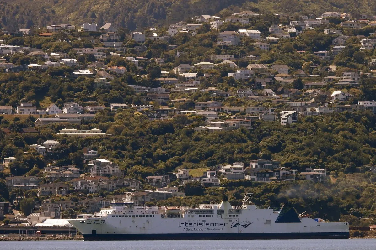 عبارة \"Interislander\" تبحر في مضيق كوك، محاطة بمنازل خضراء على تل، تعكس تأثير الطقس القاسي على السفر في نيوزيلندا.